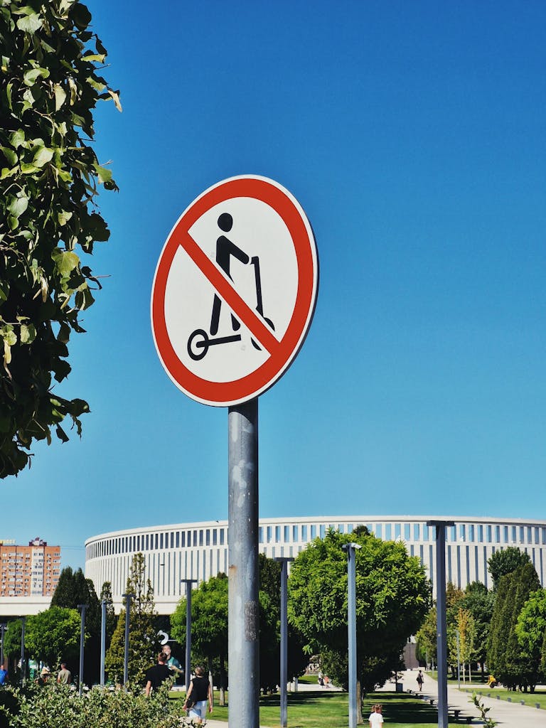 A no scooter sign in a sunny urban park with people and trees.